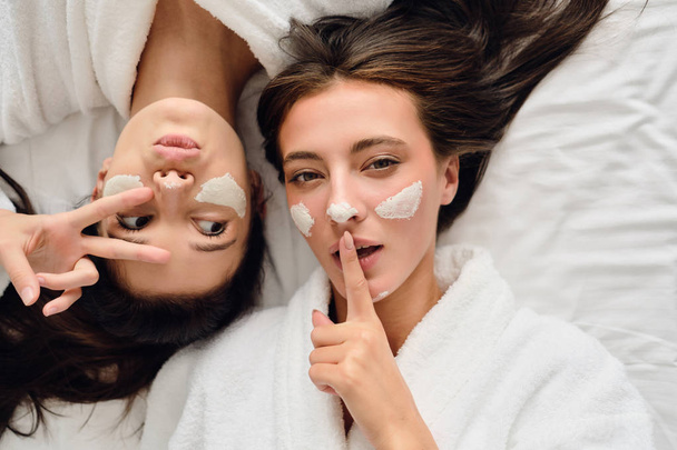 Two young attractive women with dark hair in white bathrobes lying near in bed with cosmetic mask on faces thoughtfully looking in camera together in modern cozy hotel  - Photo, Image