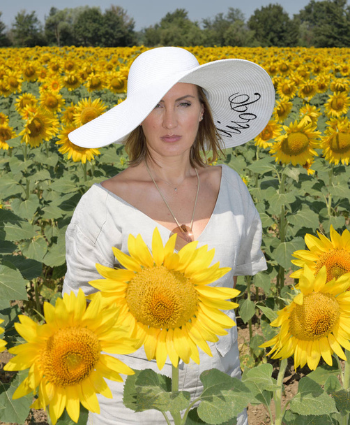 Portrait pf a young and beautiful woman looking at the camera in a sunflower field wearing an oversized hat - Photo, Image