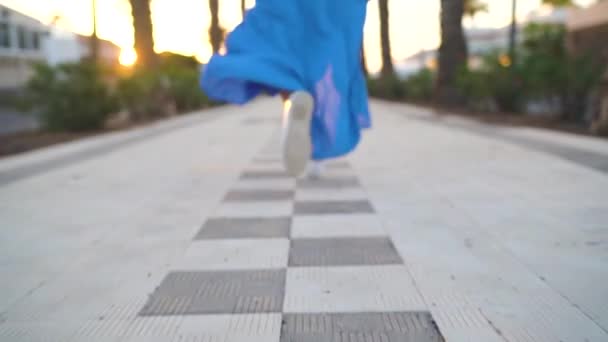 Legs of a woman in a beautiful blue dress running along the palm avenue. Hat falls to the ground - Felvétel, videó