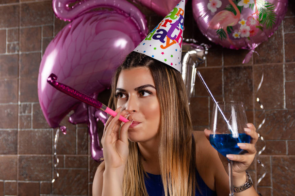 Boldog fiatal nőt zenei blowouts és koktél kék lagúna a kezében, és sapkát a fejét ünnepli születésnapját egy éjszakai klubban. Születésnapi party koncepció. Közelről, szelektív fókusz - Fotó, kép