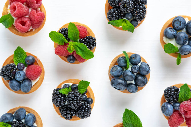 Photo pattern with tartlets on a white wooden background. Cakes with berries of marina, blueberry, blueberry with mint leaves. Photo patern with sweets top view - Photo, image