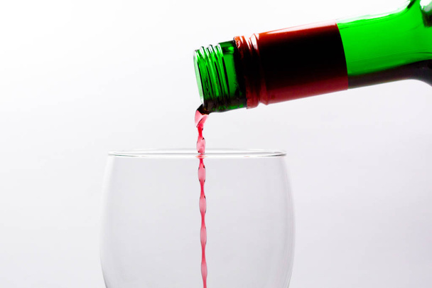 Γεμίζοντας ένα ποτήρι ποτό με κόκκινο κρασί, από το πράσινο μπουκάλι ένα πράσινο μπουκάλι. Σε λευκό φόντο. - Φωτογραφία, εικόνα