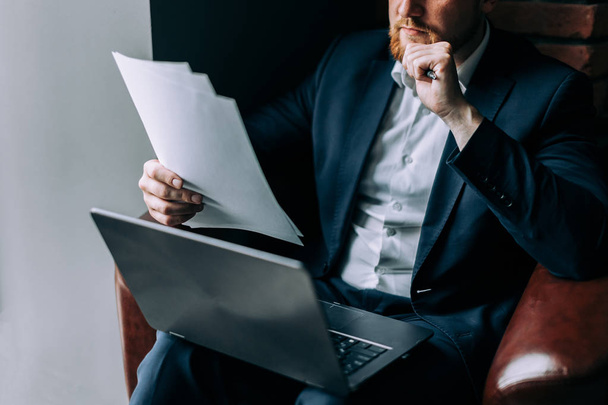 Un uomo d'affari in giacca e cravatta si siede su una poltrona con un computer portatile e analizza le informazioni di un giornale
. - Foto, immagini