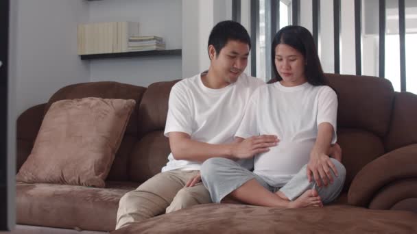 Young Asian Pregnant couple man touch his wife belly talking with his child. Mom and Dad feeling happy smile peaceful while take care baby, pregnancy lying on sofa in living room at home. Slow motion. - Filmati, video