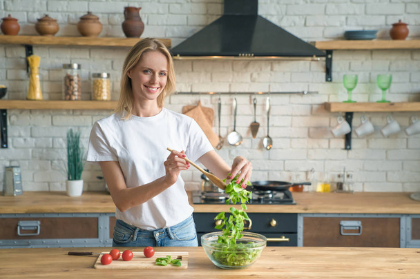 hermosa joven sonriente mujer haciendo ensalada en la cocina. Comida saludable. ensalada de verduras. Dieta. Estilo de vida saludable. cocinar en casa. Usar camisa blanca y jeans
. - Foto, imagen