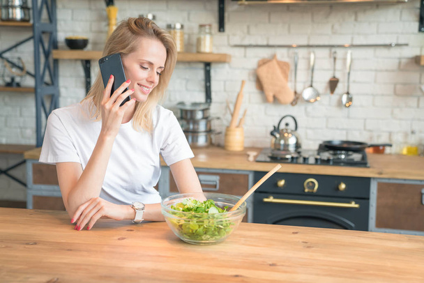 beautiful young smiling woman talking on the phone while making salad in the kitchen. Healthy food. vegetable salad. Diet. Healthy lifestyle. cooking at home. Wearing white shirt. - Photo, Image