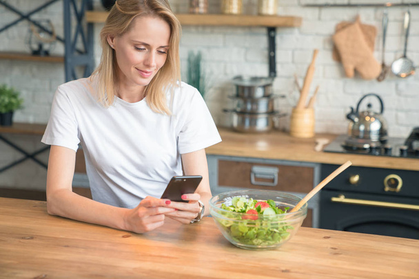 красивая молодая женщина с помощью мобильного телефона, делая салат на кухне. Здоровое питание. овощной салат. Диета. Здоровый образ жизни. приготовление пищи дома. В белой рубашке и джинсах
. - Фото, изображение