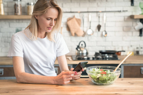 beautiful young woman using cell phone while making salad in the kitchen. Healthy food. vegetable salad. Diet. Healthy lifestyle. cooking at home. Wearing white shirt and jeans. - Фото, зображення