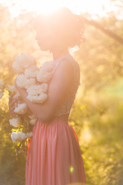 Jovem atraente em um vestido longo com flores brancas em suas mãos em um parque de verão ao pôr do sol
 - Foto, Imagem