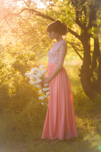 Attractive young girl in a long dress with white flowers in her hands in a summer park at sunset - Φωτογραφία, εικόνα