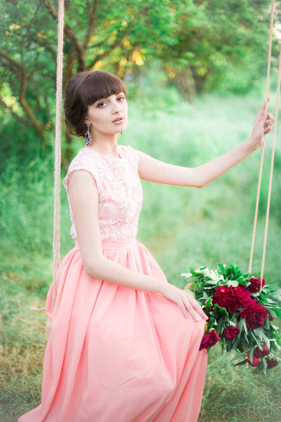 Attractive young girl in a long dress, with a bouquet of burgundy peonies in her hands on a swing in a summer park at sunset - Фото, зображення