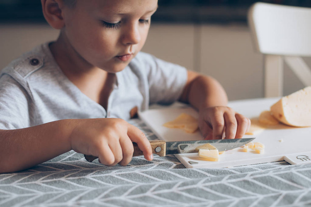 Cute boy 4-5 years old with knife cutting a cheese on the cutting board on the table in the kitchen. Dairy product. Healthy eating and lifestyle. - Photo, Image