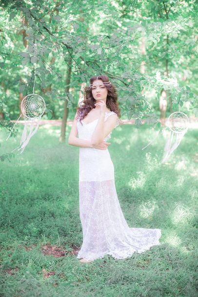 Jong aantrekkelijk meisje in een lange witte jurk met een mooi kapsel, met witte droom vangers in een zomerpark - Foto, afbeelding