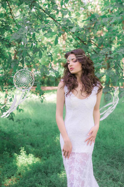 Young attractive girl in a long white dress with a beautiful hairstyle, with white dream catchers in a summer park - Foto, Bild