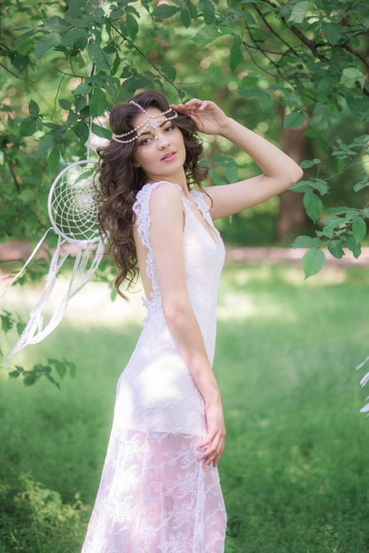 Young attractive girl in a long white dress with a beautiful hairstyle, with white dream catchers in a summer park - Foto, immagini