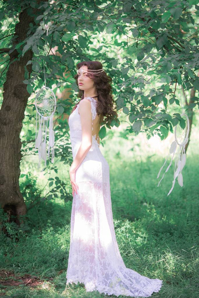 Young attractive girl in a long white dress with a beautiful hairstyle, with white dream catchers in a summer park - Foto, Imagen