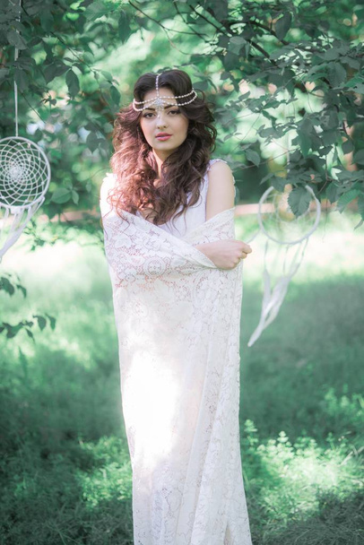 Attractive young girl in a long white dress with a beautiful hairstyle, with a white lace vintage scarf in a summer park - Foto, Imagem