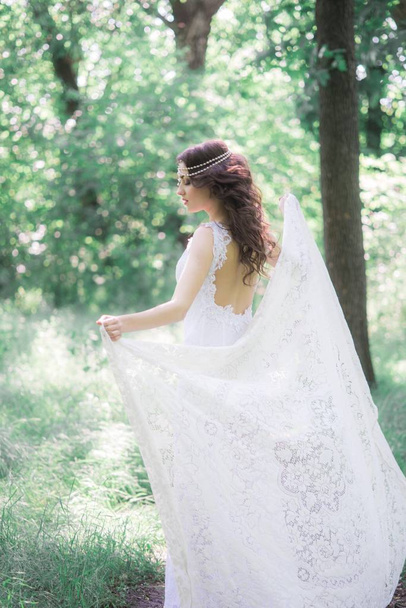 Attractive young girl in a long white dress with a beautiful hairstyle, with a white lace vintage scarf in a summer park - Photo, Image
