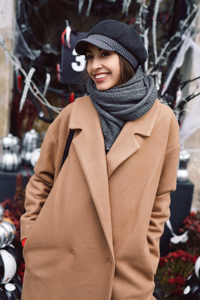 Autumn lifestyle portrait of beautiful smiling woman in beige coat, warm scarf and hat on the street with halloween background. - Valokuva, kuva