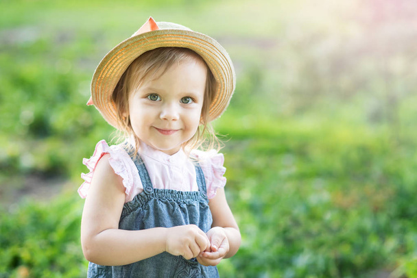 little farmer girl in hat smiling and looking at camera while posing in summer garden - Photo, Image