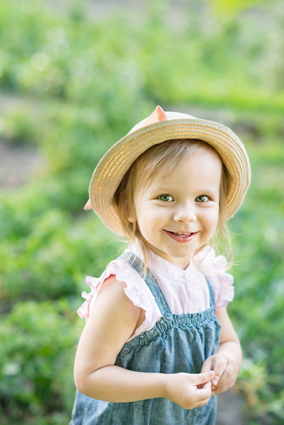 little farmer girl in hat smiling and looking at camera while posing in summer garden  - Fotó, kép