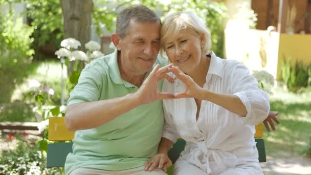 Elderly couple hands show heart. Elderly couple sitting on a bench in an embrace. Man and woman sitting in the garden and smile. - Footage, Video