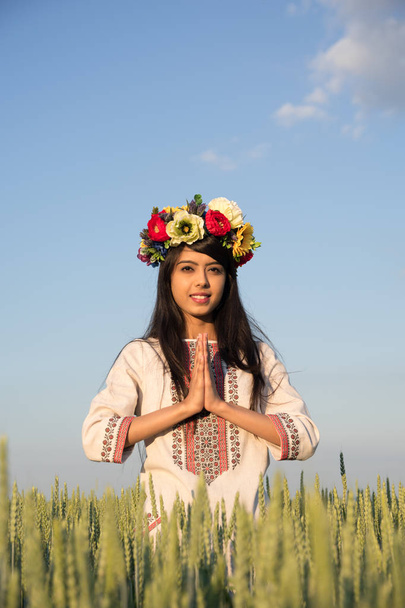 Hermosa chica india sonriente en ropa tradicional ucraniana y flores hechas a mano corona de pie en el campo de centeno con las manos en gesto de saludo namaste, mujer joven étnica en el cielo fondo de la naturaleza
 - Foto, imagen