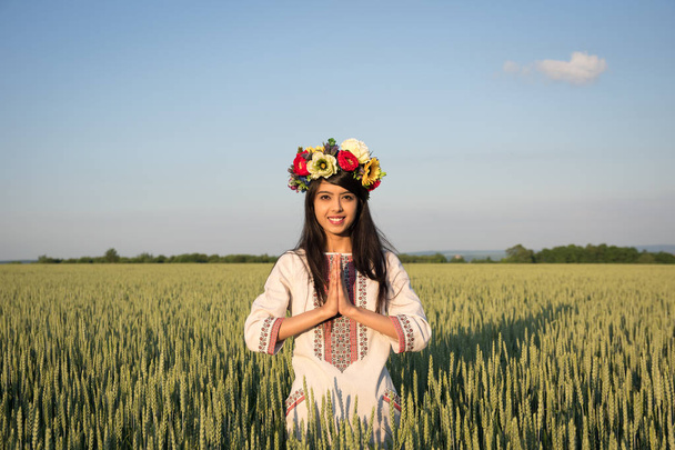 Geleneksel Ukrayna giysileri ve el yapımı çiçekli çelenk namaste tebrik jest elleri ile çavdar alanında güzel gülümseyen hintli kız, gökyüzü doğa arka planda etnik genç kadın - Fotoğraf, Görsel