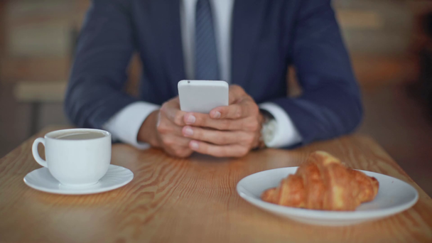 wykadrowany widok biznesmena z rogalikiem i kawą za pomocą smartfona - Materiał filmowy, wideo