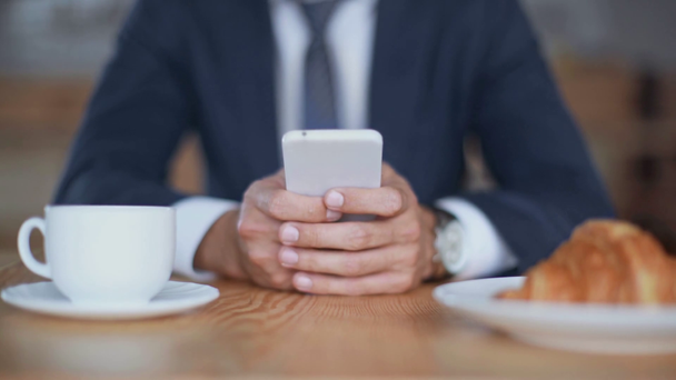 cropped view of businessman using smartphone in cafe with cup of coffee and croissant - Footage, Video