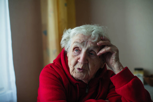 Vieja solitaria en su casa, sentada en una mesa con una chaqueta roja. La difícil situación de los pensionistas
. - Foto, imagen
