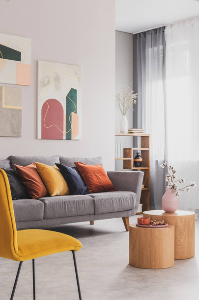 Yellow, orange, black and brown pillows on comfortable grey scandinavian sofa in bright living room interior with abstract paintings on the wall - Fotó, kép