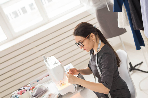 People, dressmaker and fashion concept - Side view portrait of fashion designer in glasses using sewing machine in workshop - Fotografie, Obrázek