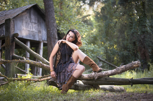 Nuori kaunis nainen istuu tukit metsässä lähellä vanhaa taloa
 - Valokuva, kuva