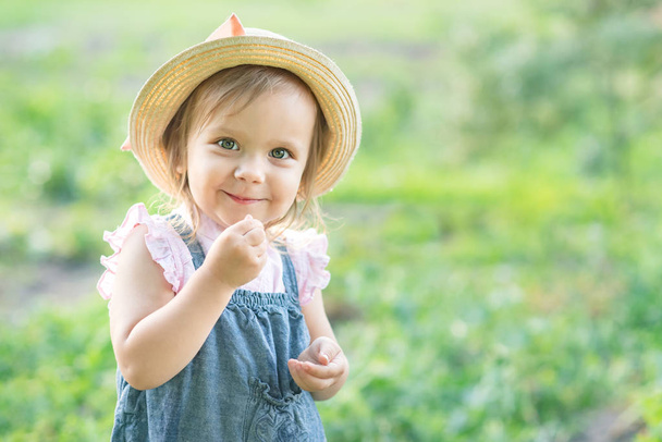 little farmer girl in hat eating pea pod in summer garden. Healthy eating for kid concept  - 写真・画像