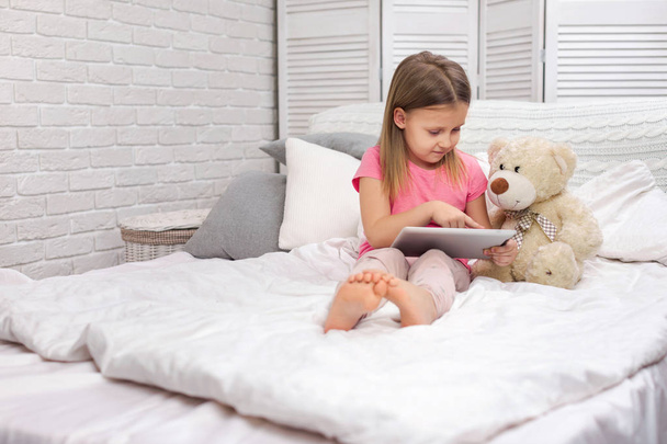 cute little girl lies in bed uses digital tablet. - Zdjęcie, obraz