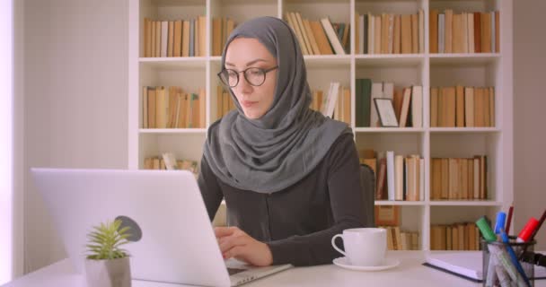 Closeup portrait of young attractive muslim businesswoman in glasses and hijab using laptop and drinking coffee in library indoors - Πλάνα, βίντεο