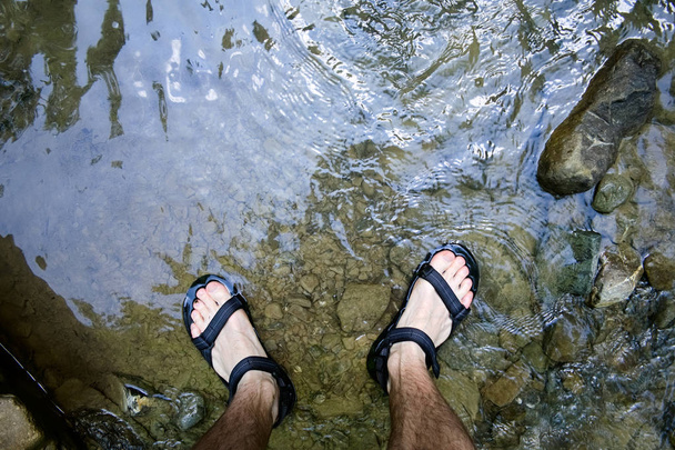 Genç adamın bacakları suya batırılmış. Kayaların üzerindeki Soğuk Dağ Nehri'nde sandalet ayakkabılarını giyen gezginler. Karpat dağları, Ukrayna. Seçici odaklama. - Fotoğraf, Görsel