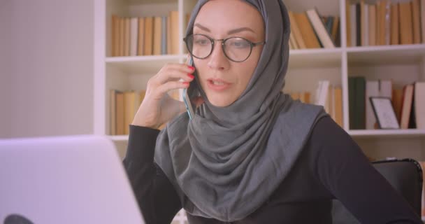 Zbliżenie Portret młodej ruchliwej muzułmańskiej bizneswoman w hidżab za pomocą laptopa i o rozmowę telefoniczną notatek w bibliotece w pomieszczeniu - Materiał filmowy, wideo