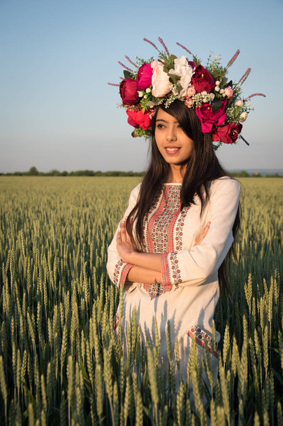 Beautiful indian young woman in traditional Ukrainian embroidery clothes and floral handmade wreath stand in field, look in distance dreaming or thinking, pretty ethnic woman posing in nature  - Photo, Image