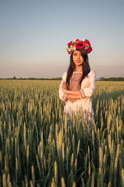 Schöne indische junge Frau in traditioneller ukrainischer Stickkleidung und floralem handgefertigten Kranz stehen im Feld, schauen in der Ferne träumend oder denkend, hübsche ethnische Frau posiert in der Natur  - Foto, Bild