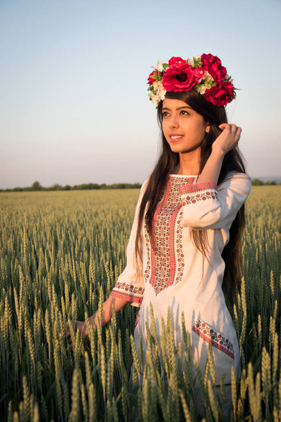 ウクライナの刺繍でポーズを取るウクライナの刺繍のインドの女性伝統的なウクライナの刺繍服と花の手作りの花輪のスタンドでフィールドでポーズをとるインドの女性は、遠くに夢や思考、自然の中でポーズのかわいい民族の女性を見て  - 写真・画像