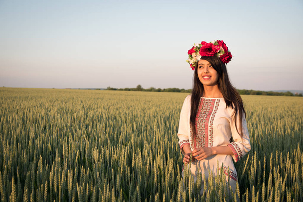Indian woman in Ukrainian embroidery posing in fieldBeautiful indian young woman in traditional Ukrainian embroidery clothes and floral handmade wreath stand in field, look in distance dreaming or thinking, pretty ethnic woman posing in nature  - Photo, Image