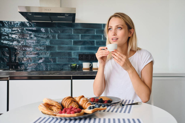 junge attraktive blonde Frau in weißem T-Shirt verträumt mit einer Tasse Kaffee in der Hand am Tisch mit Frühstück im modernen gemütlichen Zuhause - Foto, Bild