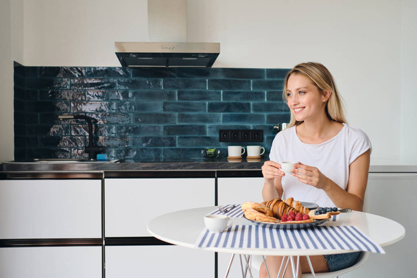 Joven hermosa mujer rubia sonriente en camiseta blanca felizmente mirando a un lado con una taza de café en la mano sentada en la mesa con el desayuno en el hogar moderno
 - Foto, imagen