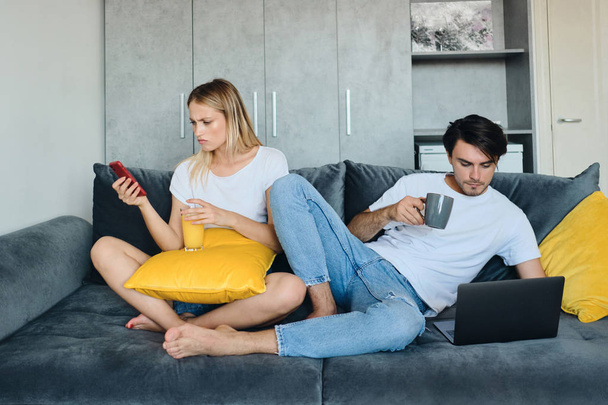 Die aufgebrachte blonde Frau mit einem Glas Orangensaft in der Hand bedient das Handy, während der nachdenkliche brünette Mann mit einer Tasse Kaffee am Laptop arbeitet. junges Paar sitzt auf Sofa im modernen Zuhause - Foto, Bild
