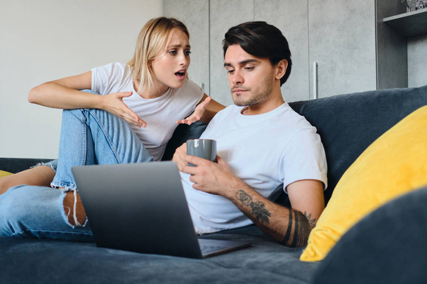 Sad emotional blond woman angrily talking to calm brunette man with cup of coffee in hands and laptop near. Young couple in bad mood sitting on sofa in modern home - Foto, afbeelding