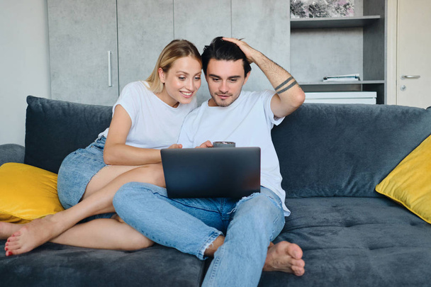 Pretty smiling blond woman and attractive brunette man happily using laptop together. Young beautiful couple sitting on sofa at modern cozy home - Fotó, kép