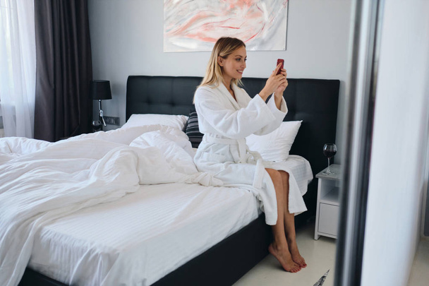 Mladá krásná usměvavá blondýnka v bílém županu s radostí používá mobilní telefon sedící na velké posteli v moderním útulném hotelu - Fotografie, Obrázek