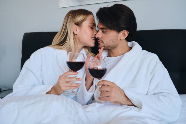 Νέος μελαχρινή άντρας και όμορφη ξανθιά γυναίκα σε λευκά μπουρνούζια ονειρικά φιλιά με γυαλιά κόκκινου κρασιού στα χέρια. Όμορφο ζευγάρι που βρίσκεται στο κρεβάτι στο μοντέρνο φιλόξενο ξενοδοχείο - Φωτογραφία, εικόνα
